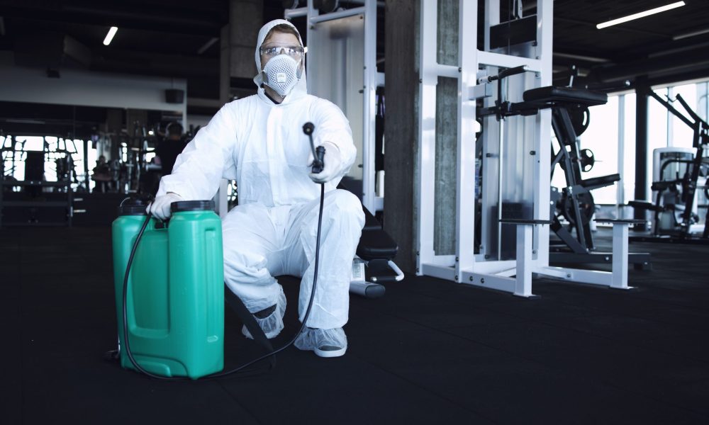 Man in white protection suit disinfecting and fitness equipment and weights to stop spreading highly contagious corona virus.